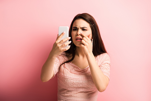nespokojená dívka dotýkající se nosu při pohledu na smartphone na růžovém pozadí - Fotografie, Obrázek