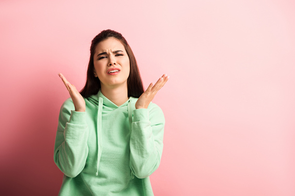 upset girl looking at camera while standing with open arms on pink background - Foto, Imagem