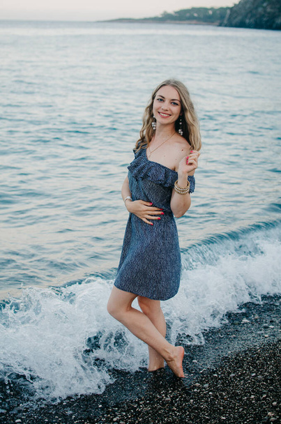 Ρομαντικό ταξιδιώτη γυναίκα σε ένα μπλε φόρεμα στην ακτή της Μεσογείου θάλασσας. Το μοντέλο στέκεται ξυπόλητο στα κύματα, σκίζοντας τον ώμο της, ποζάροντας και χαμογελώντας. Απολαμβάνει τις διακοπές στην παραλία. Αντιγραφή χώρου - Φωτογραφία, εικόνα