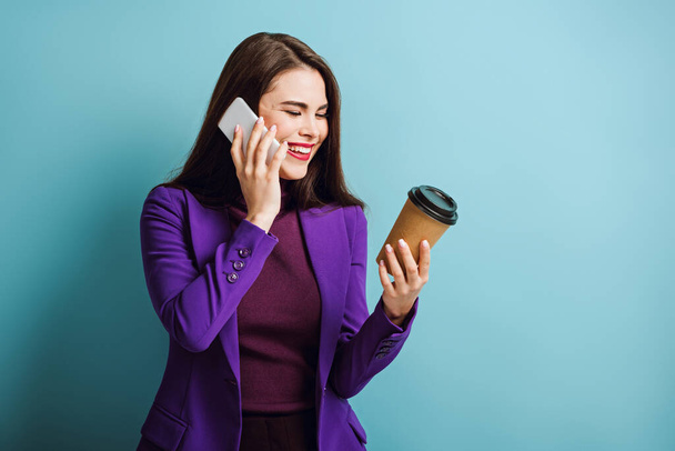 vrolijk jong vrouw praten op smartphone terwijl het houden van koffie te gaan op blauwe achtergrond - Foto, afbeelding