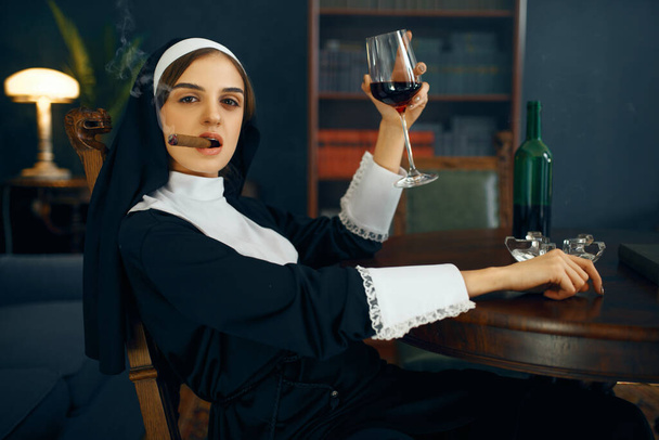 Sexy nun in a cassock smokes cigare and drinks wine, vicious desires. Corrupt sister in the monastery, religion and faith, sinful religious people, attractive sinner - Photo, Image