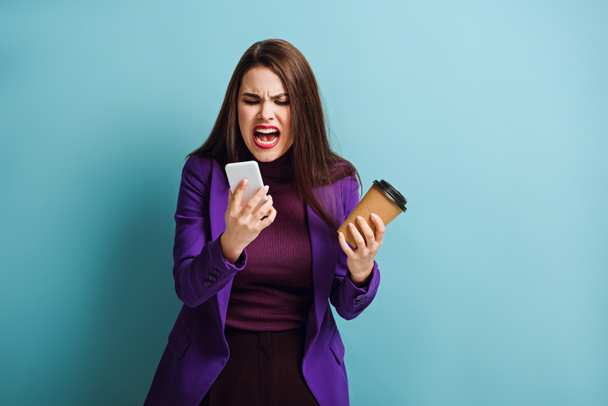 сердита дівчина тримає каву, щоб піти і кричить під час відеодзвінка на смартфон на синьому фоні
 - Фото, зображення