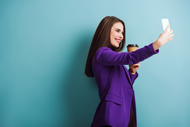 cheerful girl taking selfie on smartphone while holding coffee to go on blue background - Fotografie, Obrázek
