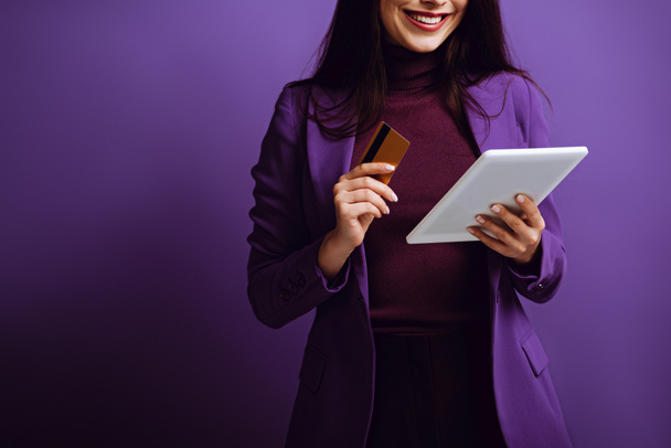 oříznutý pohled usměvavé ženy držící digitální tablet a kreditní kartu na fialovém pozadí - Fotografie, Obrázek
