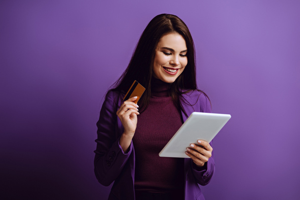 Gülümseyen genç bir kadın mor arka planda kredi kartını tutarken dijital tablete bakıyor. - Fotoğraf, Görsel
