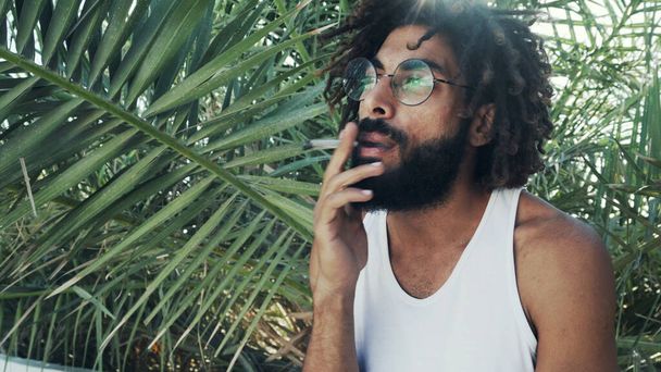 Black man with dreadlocks smoking weed joint on background of palm leafs - Photo, Image