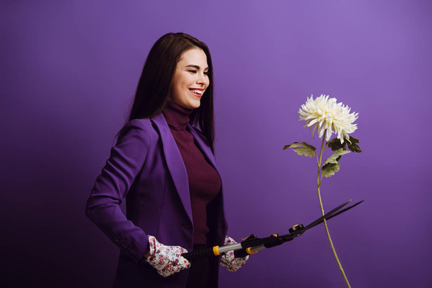 veselá dívka v textilních rukavicích řezání chryzantémy s zahradnické nůžky na fialovém pozadí - Fotografie, Obrázek