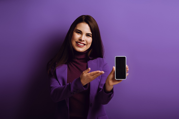 alegre joven mujer señalando con la mano en el teléfono inteligente con pantalla en blanco sobre fondo púrpura
 - Foto, imagen
