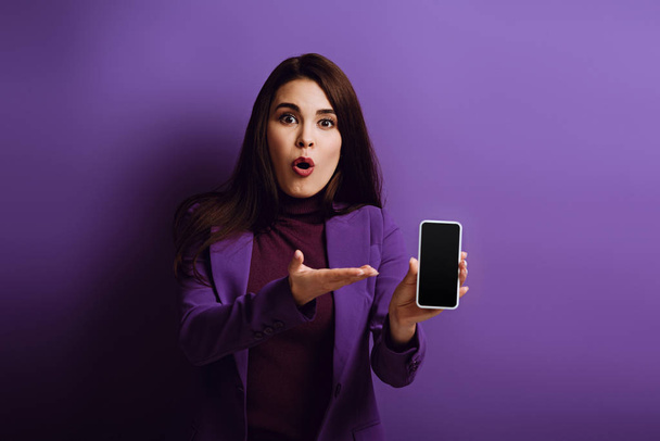 shocked girl pointing with hand at smartphone with blank screen on purple background - Photo, Image
