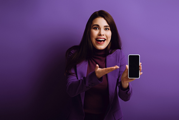 surprised girl pointing with hand at smartphone with blank screen on purple background - Fotografie, Obrázek