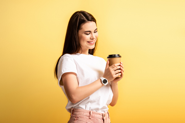 happy attractive girl smiling while holding coffee to go isolated on yellow - Fotó, kép