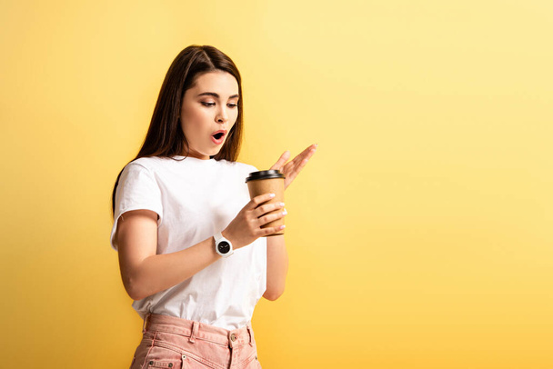 surprised girl standing with open mouth while holding coffee to go isolated on yellow - Photo, image
