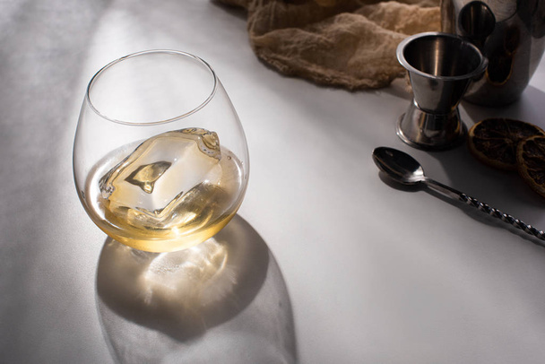 transparant glas met ijsblokje en whisky op witte tafel met schaduw bij doek, lepel en jigger - Foto, afbeelding