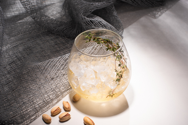 transparant glas met kruid, ijsblokje en whisky op witte tafel met schaduw bij doek en pistache - Foto, afbeelding