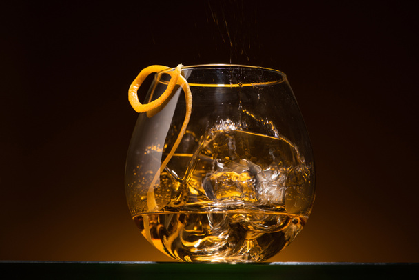 transparent glass with citrus peel, ice cubes and vodka in dark with warm back light - Photo, Image