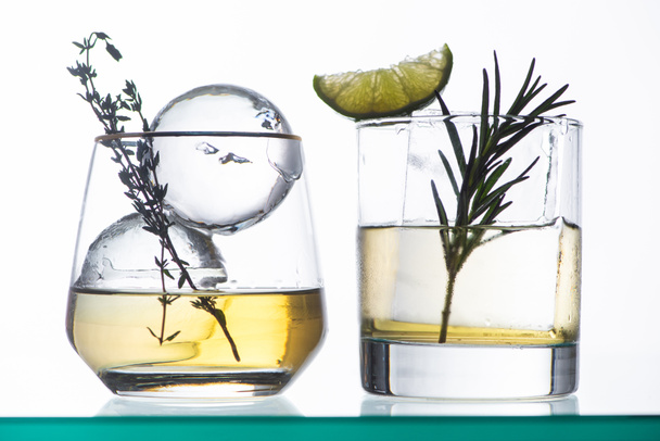 vidros transparentes com líquido dourado, ervas e gelo isolados em branco
 - Foto, Imagem