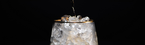 transparant glas met ijs en het gieten van gouden vloeistof geïsoleerd op zwart, panoramisch schot - Foto, afbeelding