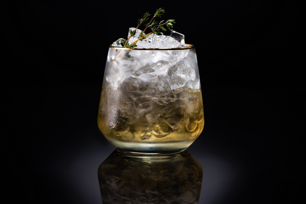 transparent glass with ice and golden liquid garnished with herb on black background - Фото, изображение