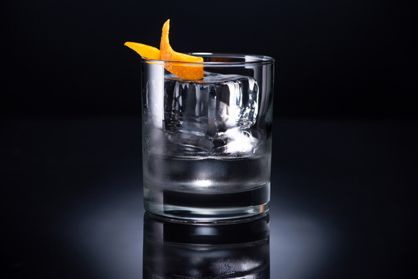 transparent glass with ice cube and vodka garnished with orange peel on black background - Foto, Bild