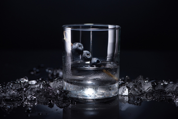 transparent glass with vodka and blueberries near smashed ice on black background - Фото, зображення