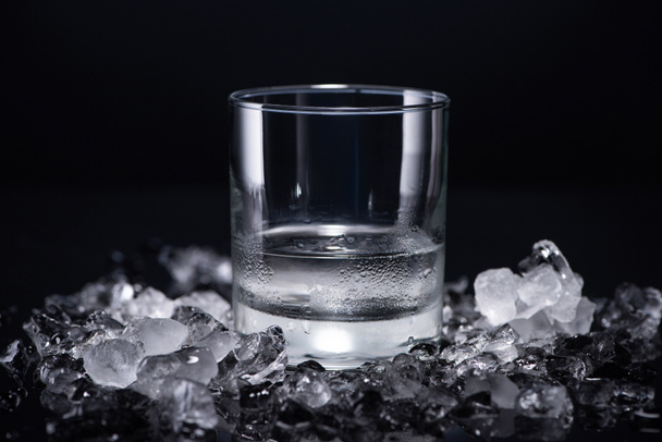 transparant glas met wodka bij ingeslagen ijs op zwarte achtergrond - Foto, afbeelding