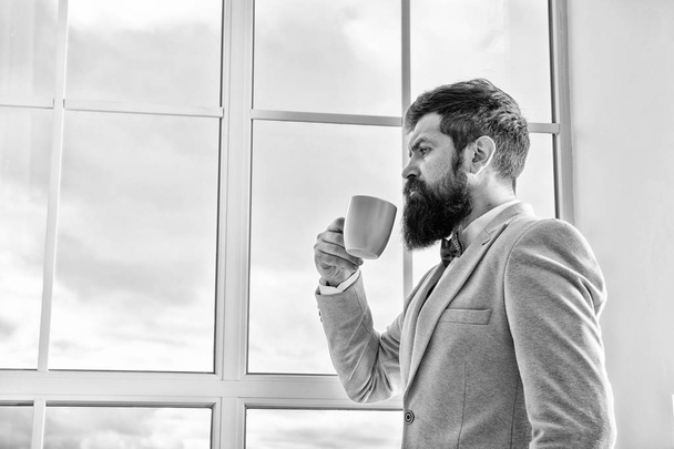 Man stand at window nervous about wedding. Life choices and expectations. But first coffee. His big day. Wedding morning concept. Wedding day. Groom bearded hipster man wear blue tuxedo and bow tie - Фото, изображение