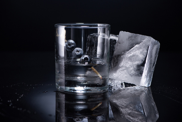 transparentní sklo s vodkou a borůvky v blízkosti kostek ledu na černém pozadí - Fotografie, Obrázek