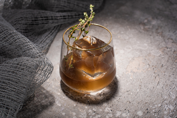 transparent glass with herb, ice cube and whiskey on concrete surface with mesh cloth - Foto, afbeelding