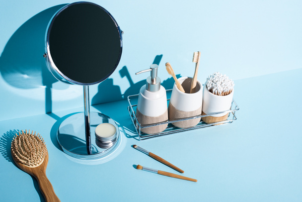 Toothbrush holders with hygiene items, dispenser on bathroom shelf with mirror, hair brush, cosmetic brushes on blue background, zero waste concept - Photo, Image