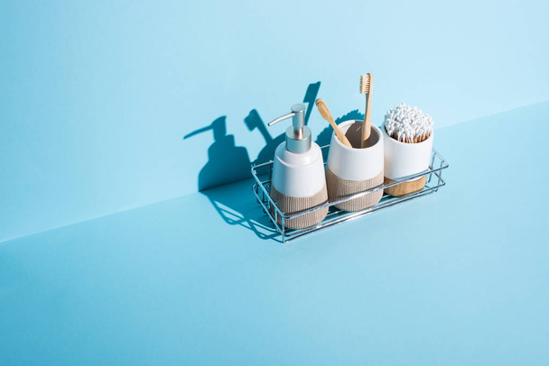Стойки для зубной щетки с палочками для ушей и зубными щетками с жидким мылом диспенсера на полке в ванной комнате на синем фоне, концепция нулевых отходов
 - Фото, изображение