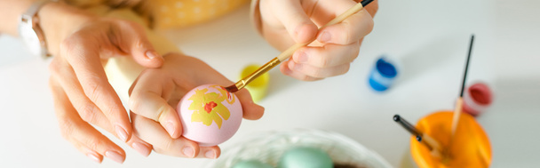 panoramic shot of kid painting easter egg near mother on white  - Foto, Bild