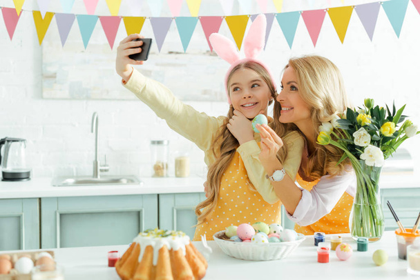 ευτυχισμένη μητέρα και κόρη στα αυτιά λαγουδάκι λήψη selfie κοντά easter κέικ και βαμμένα αυγά κοτόπουλου  - Φωτογραφία, εικόνα