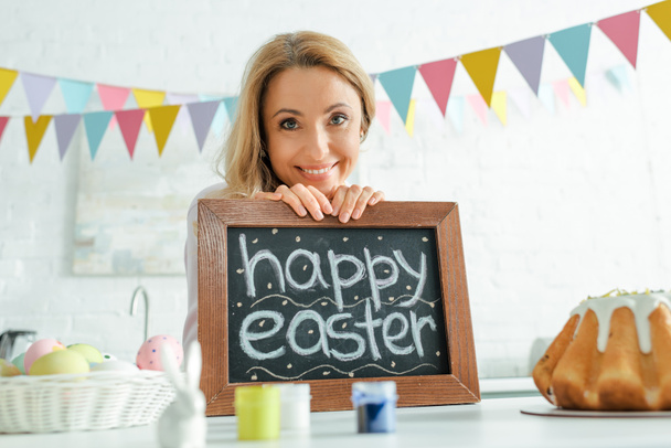 selective focus of smiling woman holding chalkboard with happy easter lettering  - Photo, Image