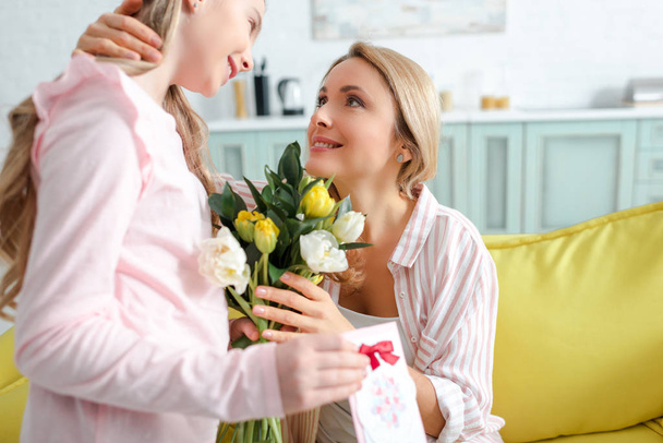selective focus of happy mother looking at daughter with flowers and greeting card  - Foto, Imagen