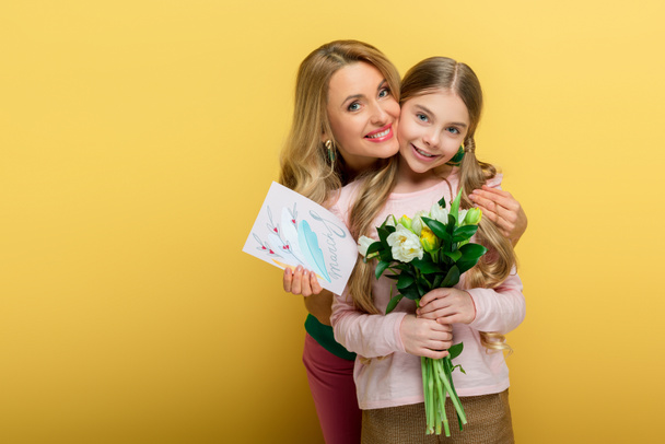 happy mother holding greeting card with 8 march lettering and hugging daughter with tulips isolated on yellow  - Foto, Bild