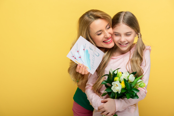 ευτυχισμένη μητέρα κρατώντας ευχετήρια κάρτα με γράμματα 8 Μαρτίου και αγκαλιάζει χαρούμενη κόρη με τουλίπες απομονωμένες στο κίτρινο  - Φωτογραφία, εικόνα