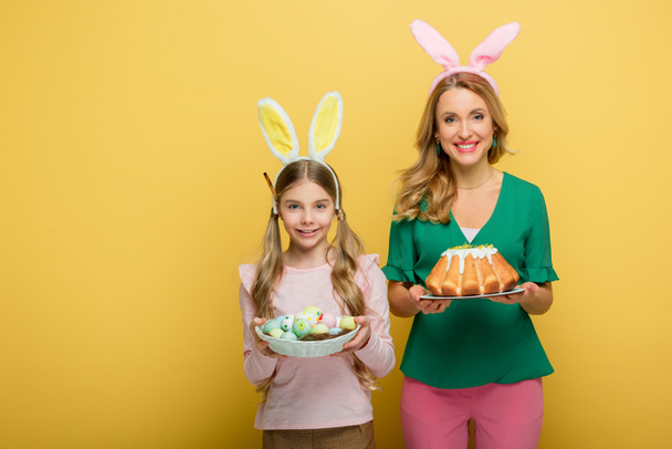 happy mother with bunny ears holding easter cake near daughter with painted chicken eggs isolated on yellow  - Фото, зображення