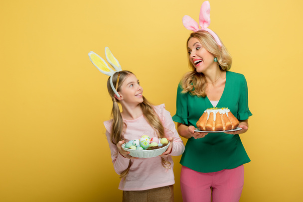 happy mother with bunny ears holding easter cake near cute daughter with painted chicken eggs isolated on yellow  - 写真・画像