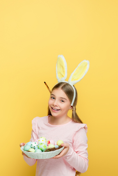 enfant heureux avec des oreilles de lapin tenant des œufs de Pâques dans le panier isolé sur jaune
  - Photo, image