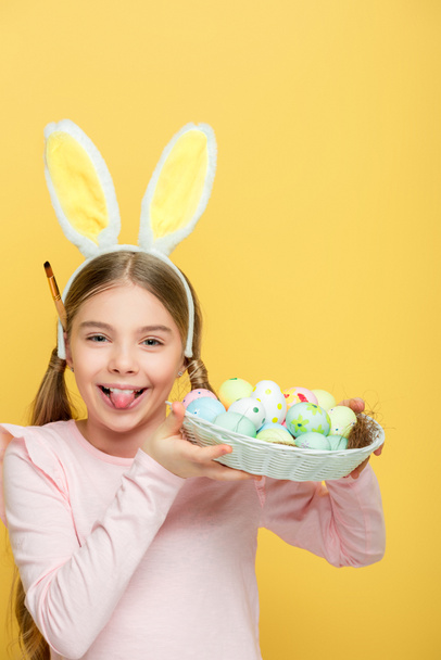 niño alegre con orejas de conejito sobresaliendo lengua y sosteniendo huevos de Pascua en cesta aislada en amarillo
  - Foto, imagen