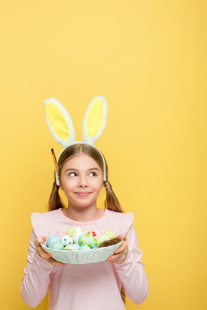 veselé dítě s králičími ušima drží velikonoční vajíčka v košíku a odvrací pohled izolované na žluté  - Fotografie, Obrázek