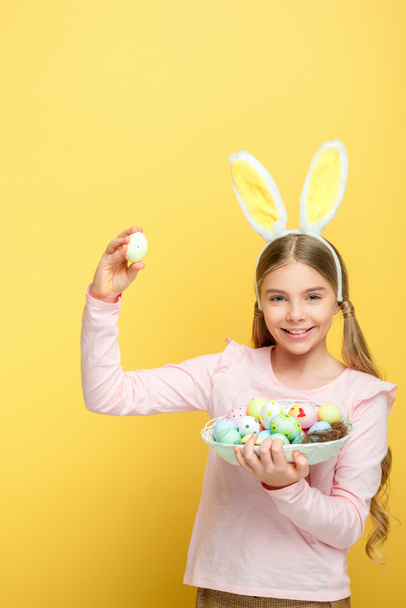 niño feliz con orejas de conejo sosteniendo huevos de Pascua en cesta aislada en amarillo
  - Foto, imagen