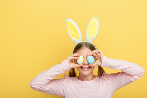 happy child with bunny ears covering eyes with easter eggs isolated on yellow  - Foto, Imagen