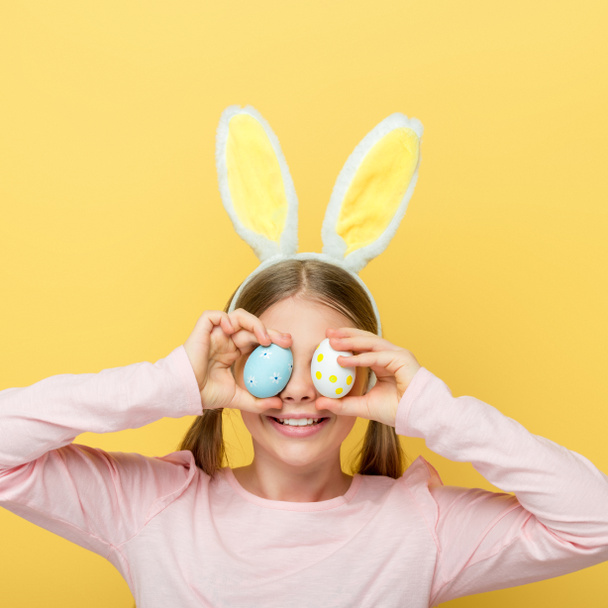 enfant gai avec des oreilles de lapin couvrant les yeux avec des œufs de Pâques isolé sur jaune
  - Photo, image