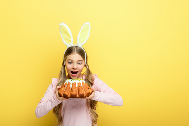 shocked kid with bunny ears holding easter cake isolated on yellow  - 写真・画像