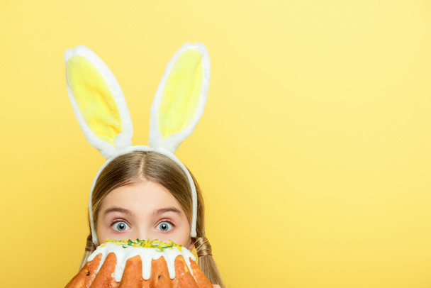 shocked kid with bunny ears covering face with easter cake isolated on yellow  - Foto, Imagem