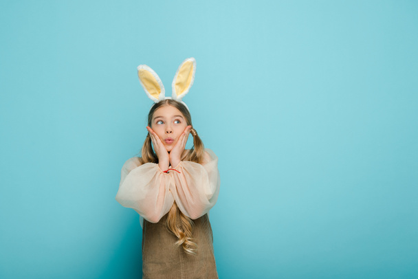 surprised kid with bunny ears touching face and looking away isolated on blue  - Photo, Image