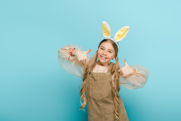 cheerful kid with bunny ears showing thumbs up isolated on blue  - Foto, Bild
