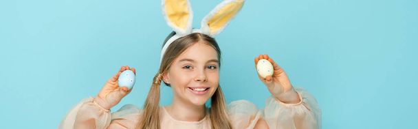 panoramic shot of happy kid with bunny ears holding painted easter eggs isolated on blue  - Photo, Image