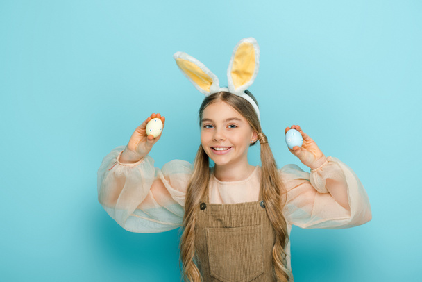 positive kid with bunny ears holding painted easter eggs on blue  - 写真・画像